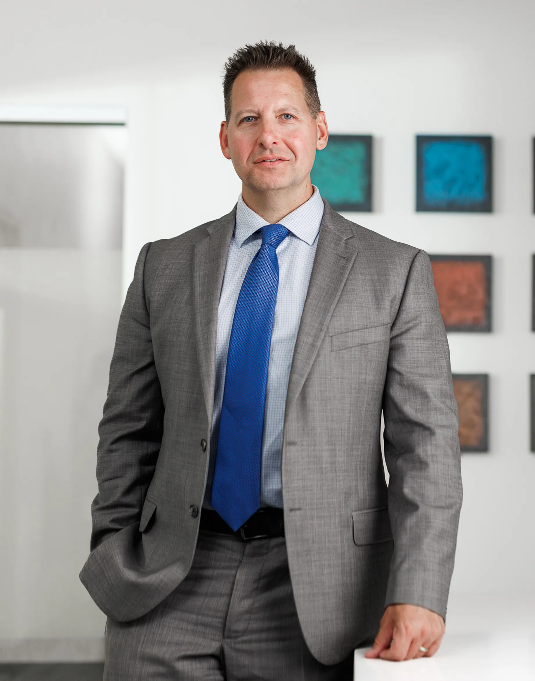 kightlinger gray michel in gray suit with blue tie on, standing in a look with frames of differnt colors on wall behind