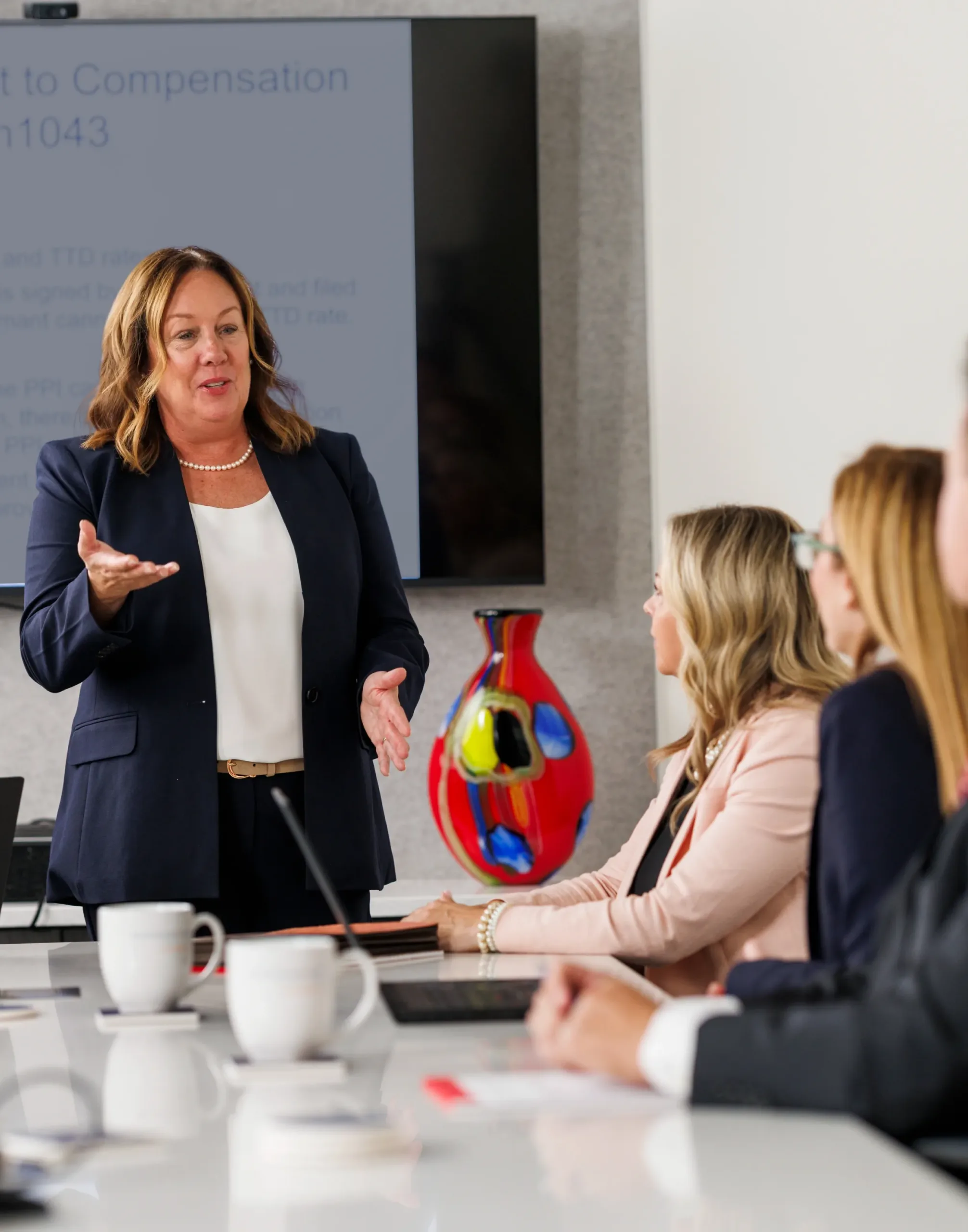 one female in blue dress giving presentation stading other 2female and one male face away hands on table half cropped