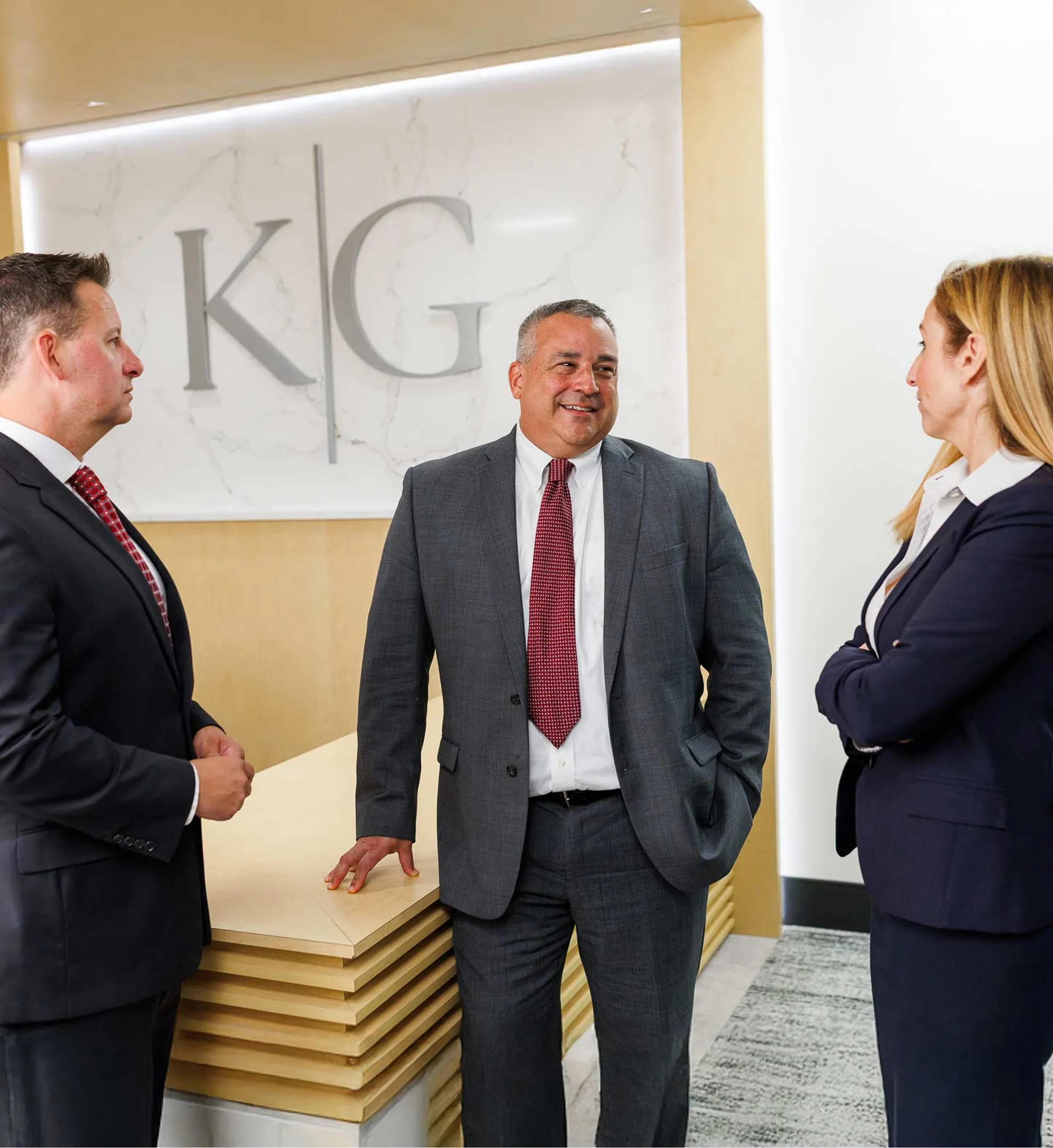 three professionals in suits one female and two males doing discussions standing in a room with K G logo on the wood art work behind on wall three professionals in suits one female and two males doing discussions standing in a room with K G logo on the wood art work behind on wall